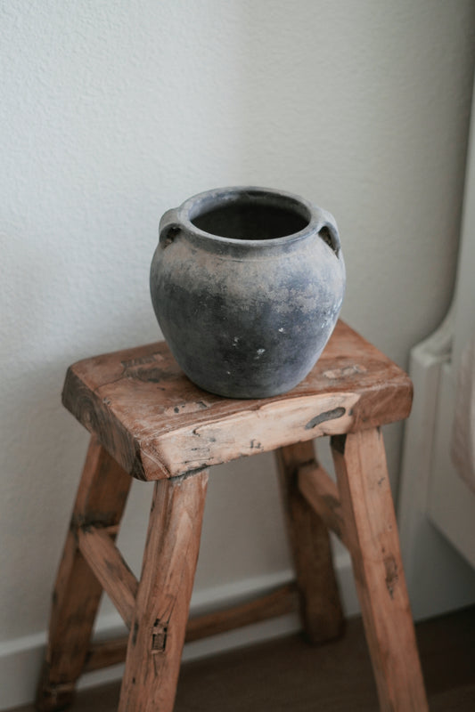 Vintage ceramic pot on a rustic wooden stool in a minimalist interior setting, showcasing a timeless, weathered design with earthy tones. Ideal for adding a touch of rustic charm and natural texture to home decor, this arrangement highlights the beauty of vintage and antique pieces in contemporary interiors. Perfect for those interested in farmhouse, wabi-sabi, or rustic home decor styles.